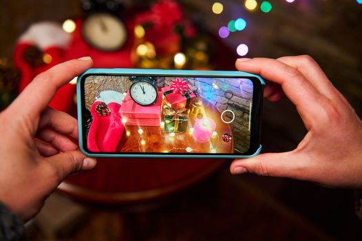 Woman hands hold smartphone, photograph a Christmas interior with luminous garland and Xmas presents