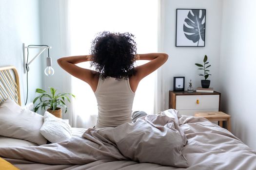 Rear view of unrecognizable woman with curly hair waking up stretching sitting on bed ledge in bedroom in the morning.