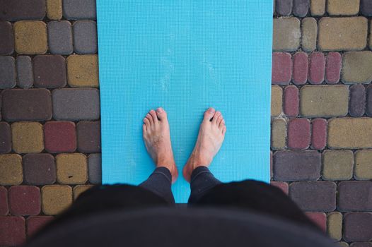 Directly above feet of active man, sportsman yogi standing barefoot on a blue fitness mat while practicing yoga outdoors