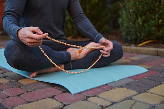 Cropped view of active male yogi sitting in lotus position, meditating with rosary, practicing yoga outdoors at sunset