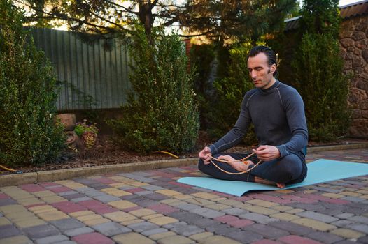 Peaceful man yogi sitting in lotus pose on a fitness mat, meditating with rosary beads outdoors at sunset. Yoga practice