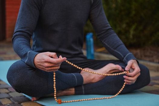 Details: hands of a yogi sitting on a fitness mat, meditating with rosary beads while practicing yoga outdoors.