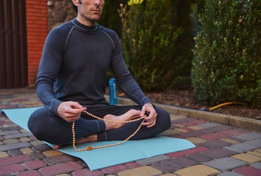 Cropped view of a yogi sitting on a fitness mat, meditating with rosary beads while practicing yoga outdoors.