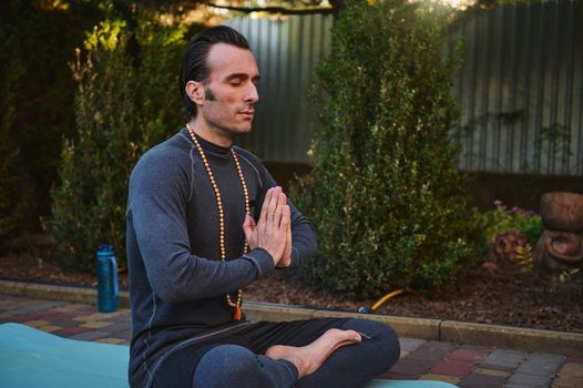 Peaceful man yogi keeping hands palms together while meditates with rosary beads in lotus pose, outdoors at sunset.