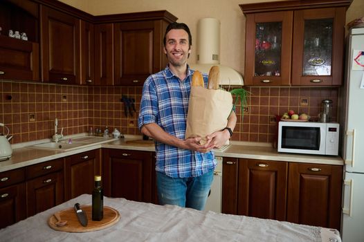 Handsome Caucasian man in blue casual shirt smiling a toothy smile at camera, holding an eco paper bag with healthy food