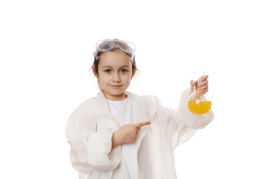 Smart school child girl points at laboratory flask with chemical solution, looks at camera, isolated on white background