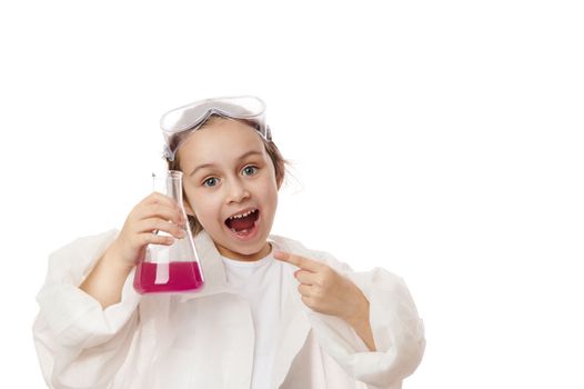 Inspired little girl points at flask with chemical liquid, smiles demonstrating the ongoing chemical reaction inside it