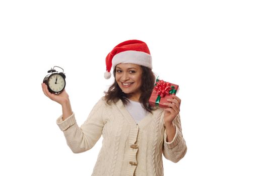 Happy multi-ethnic woman in Santa hat, holding a Xmas gift box and retro alarm clock showing five minutes to midnight