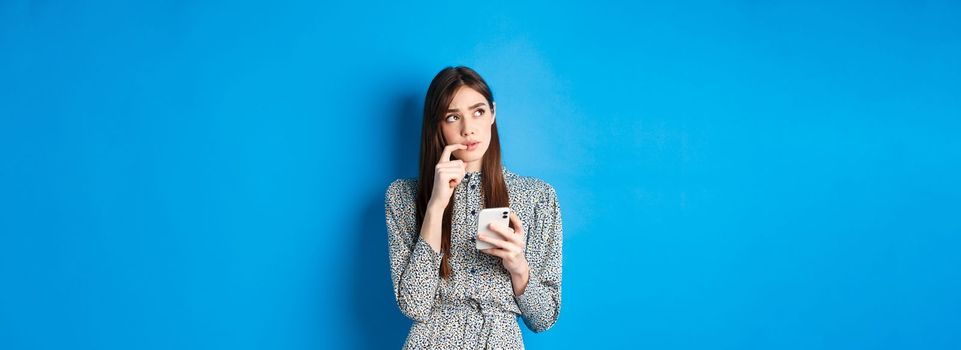 Pensive cute girl thinking how to answer on message, looking aside thoughtful and holding smartphone, standing in dress on blue background.