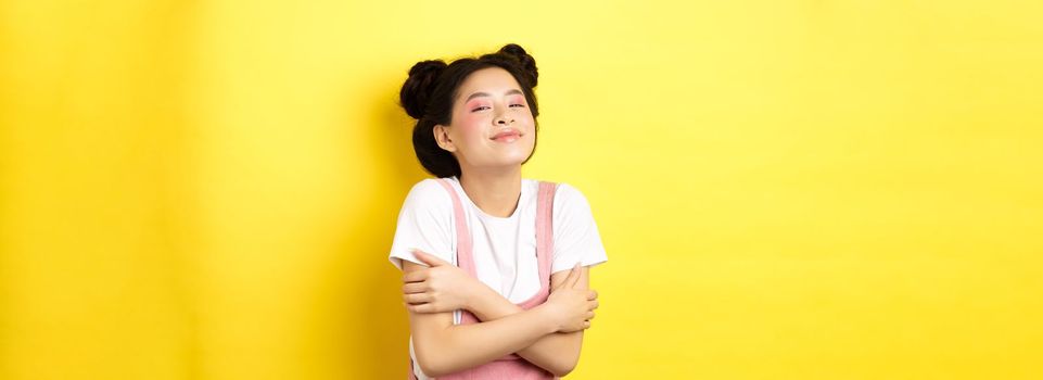 Summer lifestyle concept. Beautiful asian woman hugging herself and smiling romantic, embracing own body, standing with makeup on yellow background.