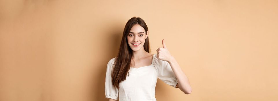 Good job. Smiling candid woman in blouse showing thumb up, praise nice choice, recommending product, standing satisfied on beige background