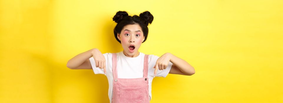 Surprised asian woman with glamour makeup and hairstyle, drop jaw and pointing down amazed, standing against yellow background