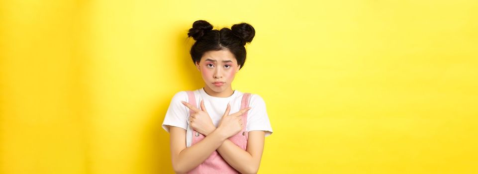 Indecisive timid asian girl pointing sideways, showing left and right ways and sulking upset, standing sad on yellow background