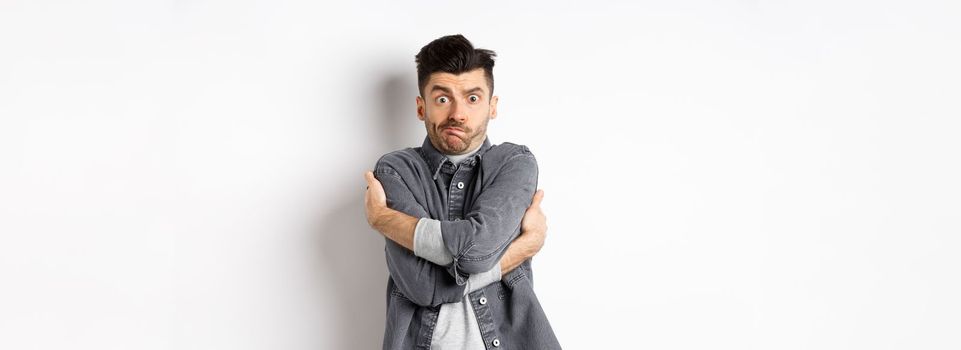 Funny young man hugging himself and grimacing, feeling embarrassed and exposed, covering body from people glances, white background