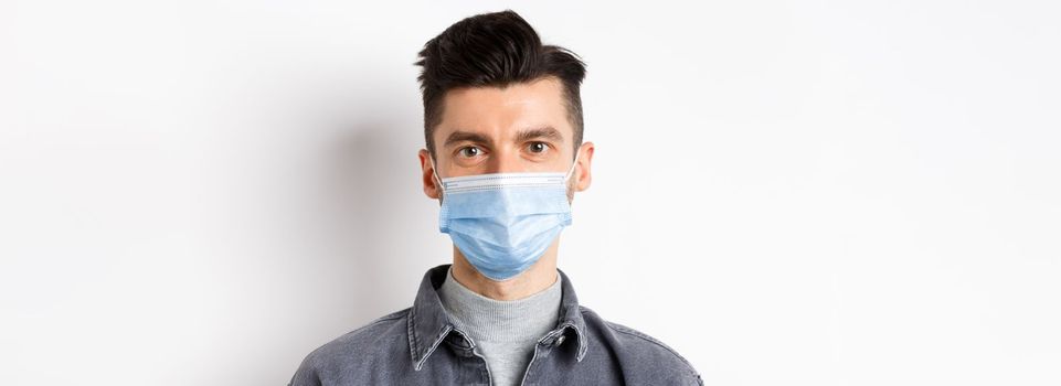 Pandemic lifestyle, healthcare and medicine concept. Close up of candid young man wearing medical mask from covid-19, taking care of health, standing on white background