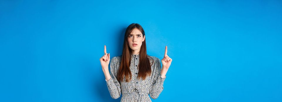 Confused attractive woman in dress frowning, pointing fingers up and looking puzzled, cant understand something, stare at strange thing, blue background