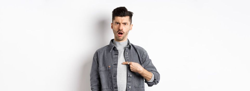 Offended young man being accused, frowning and pointing at himself shocked, standing on white background