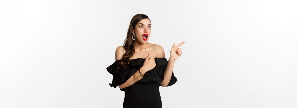 Fashion and beauty. Amazed beautiful woman in black dress open mouth fascinated, pointing fingers and looking left, white background