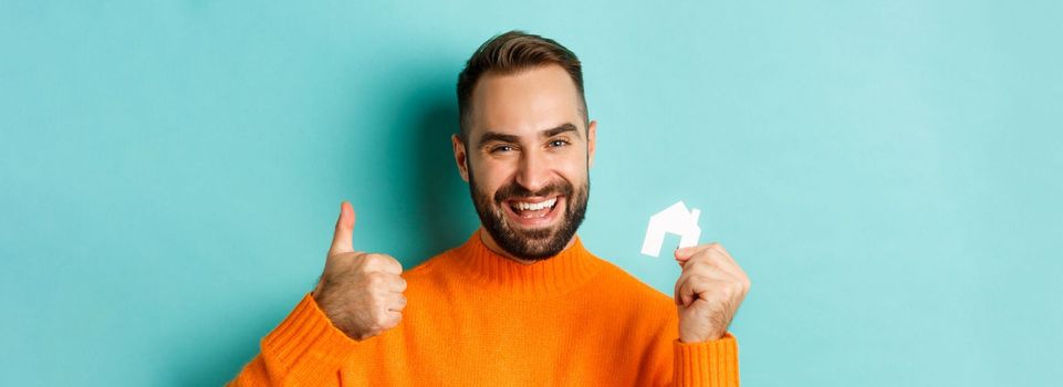 Real estate. Close-up of satisfied man showing thumb-up and small paper house maket, smiling pleased, standing over light blue background