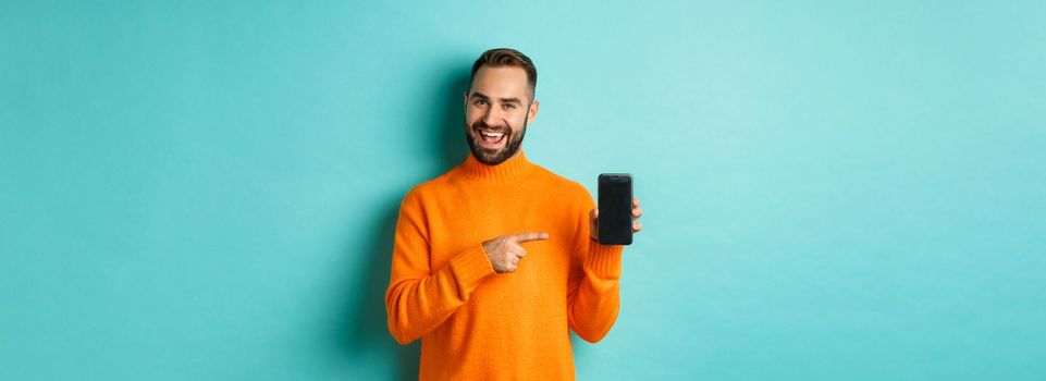Handsome bearded man in orange sweater, pointing finger at mobile phone screen, showing application smartphone, standing over light blue background