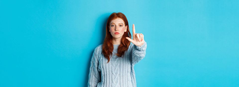 Serious redhead girl in sweater showing taboo gesture, extending one finger, shaking forefinger to disapprove, disagree and forbid something, blue background