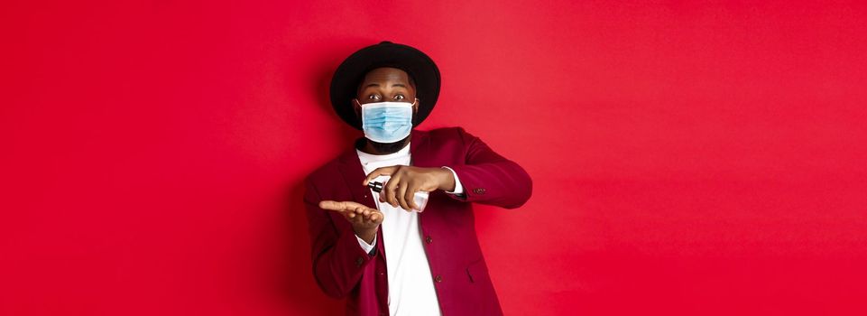 Covid-19, quarantine and holidays concept. Handsome Black man in face mask and party outfit, disinfecting hands with hand sanitizer, using antiseptic, standing over red background