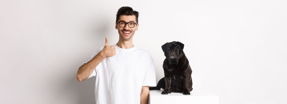 Image of dog owner and cute black pug looking at camera, man showing thumb-up in approval, recommending something, standing over white background
