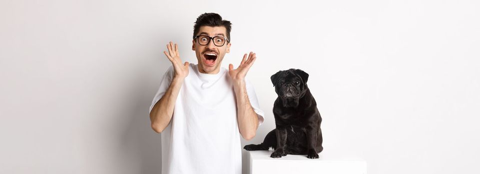 Ecstatic young man looking with excitement and rejoice, standing near cute black pug, staring at camera happy, standing over white background