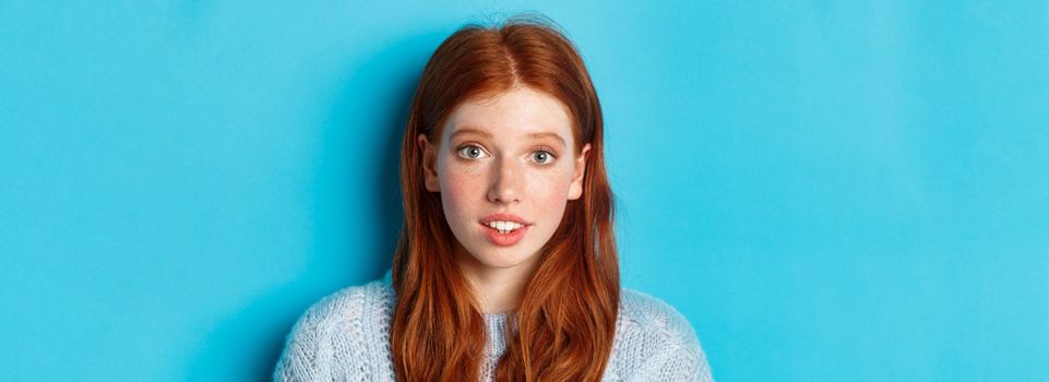 Headshot of cute redhead girl with freckles, looking hopeful and innocent at camera, standing over blue background