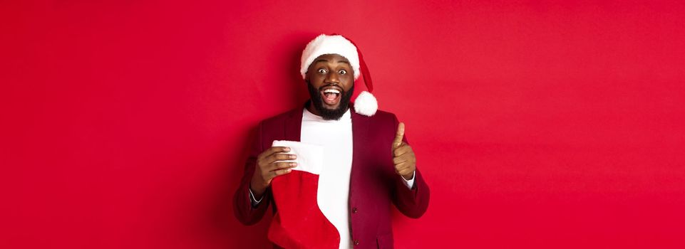 Excited Black man showing thumb-up in approval, holding christmas sock with holiday gifts, smiling amazed, standing over red background