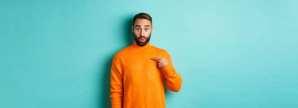 Man pointing at himself with surprise face, being chosen, standing confused against light blue background