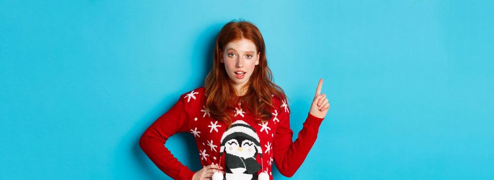 Winter holidays and Christmas Eve concept. Sassy redhead girl in xmas sweater, pointing upper left corner and staring at camera, hinting on promo offer, blue background