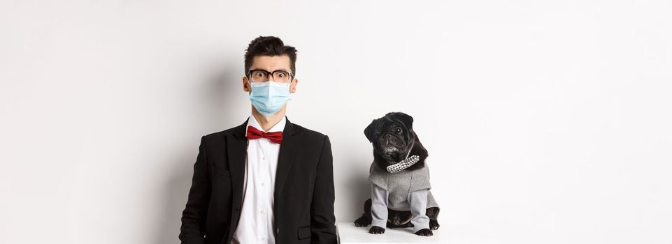 Coronavirus, pets and celebration concept. Handsome young man and dog wearing suits, guy have medical mask, standing over white background.