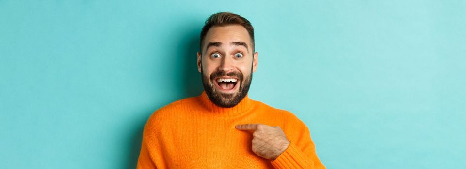 Close-up of happy young man pointing at himself with surprise and excitement, standing over light blue background