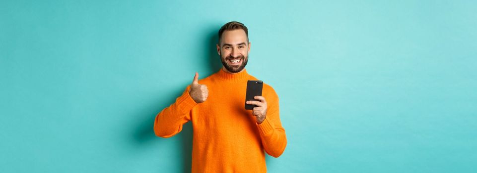 Photo of satisfied young man in orange sweater, showing thumb-up after reading on mobile phone, standing pleased against turquoise background