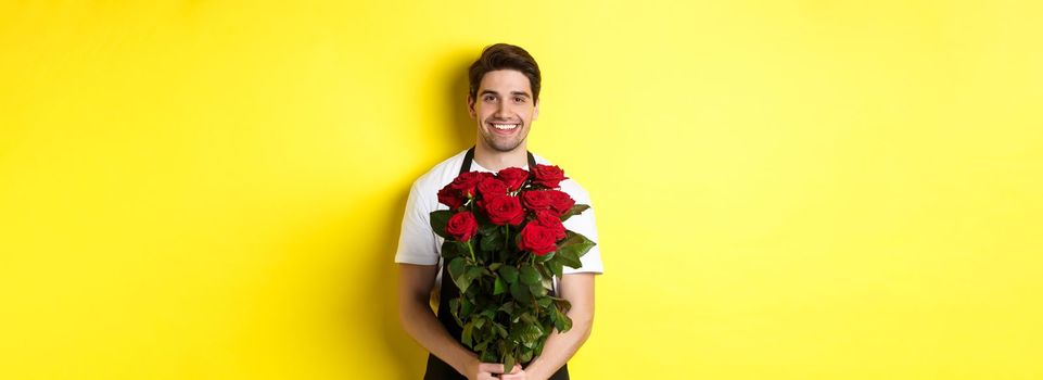 Smiling florist in black apron holding flowers, selling bouquet of roses, standing over yellow background