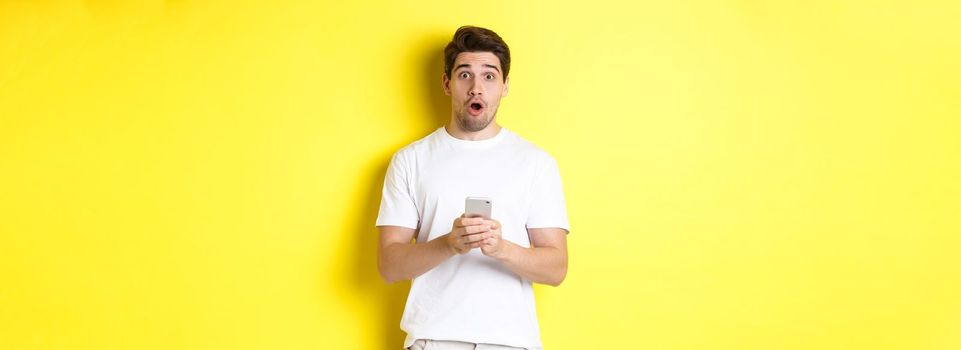 Man looking surprised, using smartphone, open mouth and saying wow, standing against yellow background