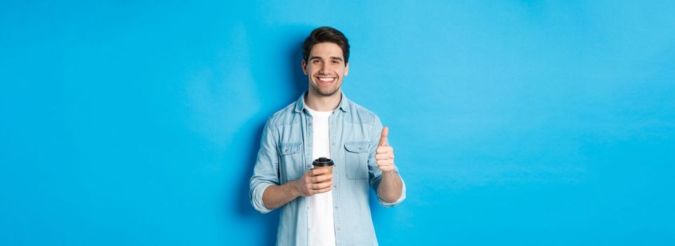 Handsome young man showing thumb-up and drinking coffee, recommending cafe takeaway, standing over blue background