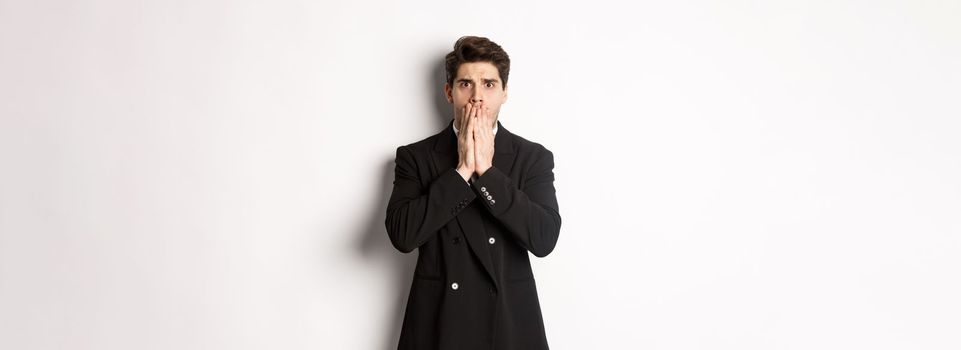 Portrait of shocked handsome businessman in suit, reacting to terrible situation, gasping and covering mouth with hands, standing startled against white background