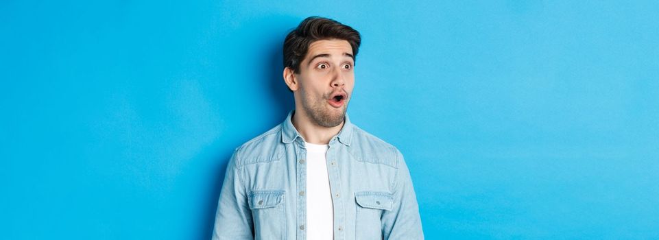 Close-up of surprised handsome man looking impressed left, open mouth fascinated, wearing casual outfit, standing over blue background