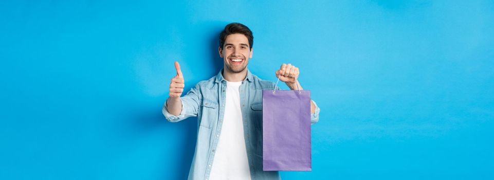 Concept of shopping, holidays and lifestyle. Satisfied smiling man holding paper bag, showing thumb-up and recommending store, standing over blue background