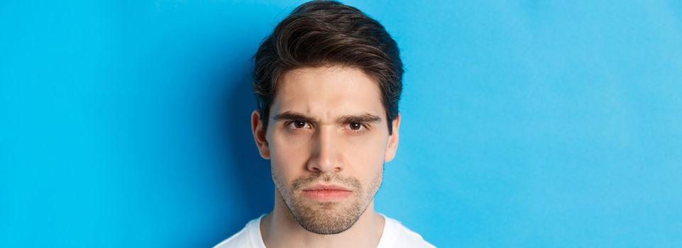 Headshot of angry man frowning, looking disappointed and bothered, standing over blue background