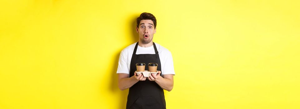 Barista serving coffee, looking surprised at camera, wearing black apron, standing against yellow background