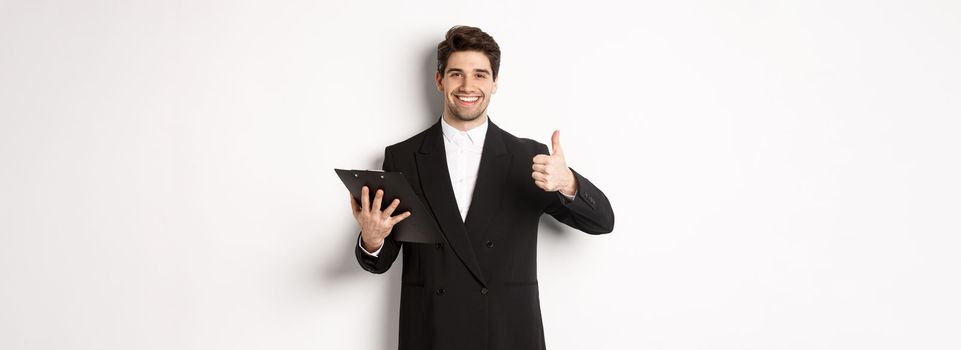 Portrait of confident businessman in black suit, holding clipboard with documents and showing thumb-up in approval, praise good job, standing against white background