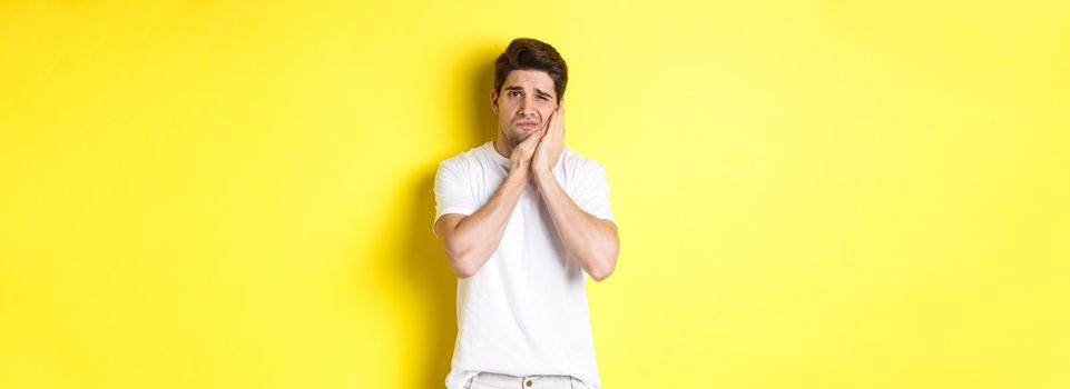 Sad guy complaining on toothache, touching swallen cheek and looking upset, standing over yellow background. Copy space