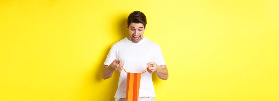 Excited guy open bag with gift, looking inside with amazement and happy face, standing against yellow background