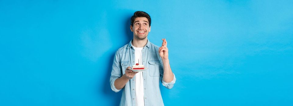 Handsome man making a wish, celebrating birthday, holding b-day cake and cross fingers, standing over blue background