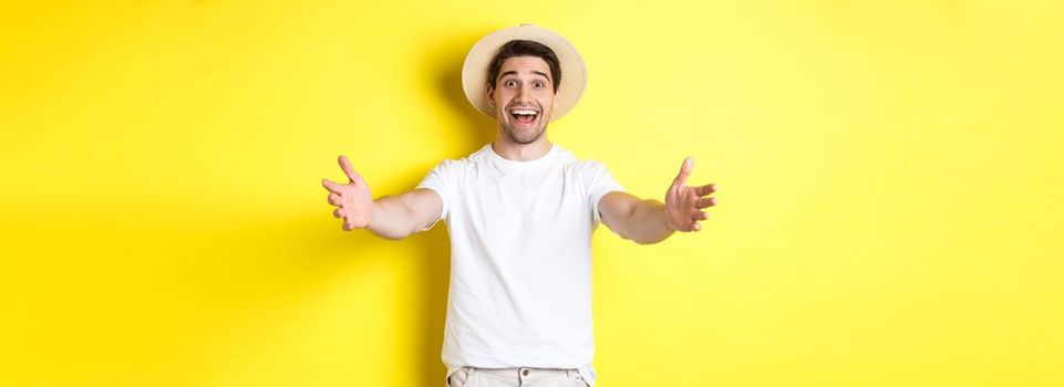 Concept of tourism and summer. Happy and friendly man traveller reaching hands for hug, greeting or welcome you, standing over yellow background