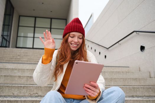 Joyful redhead teen girl, student with digital tablet, says hello, waves hand at gadget camera, connects to video chat, talks to friend in application