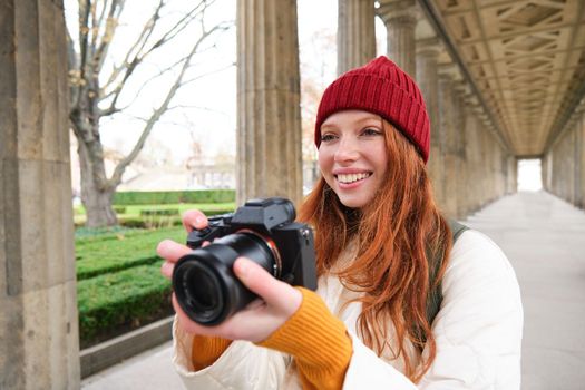 Young redhead female photographer, makes lifestyle shooting in city centre, takes photos and smiles, looks for perfect shot, makes picture